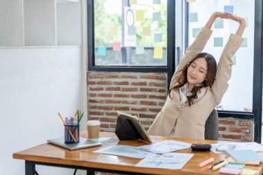 Ofis masasında otururken kollarını ve vücudunu uzatan yorgun genç bir iş kadını.