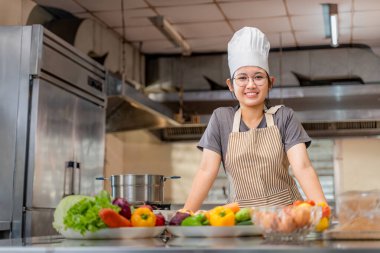 Profesyonel bir mutfakta, içi taze sebze ve yemek malzemeleriyle dolu bir tezgahın arkasında duran genç bir kadın şefin portresi. Çizgili bir önlük ve beyaz bir şef şapkası giyerek bir yemek pişirme seansına hazırlanırken kendine güvenerek gülümsüyor. Öge
