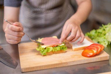 Çizgili önlük ve beyaz şef şapkası takan neşeli genç bir şef ticari bir mutfakta taze bir sandviç hazırlarken görülüyor. Bu resim, bir meslekte yemek pişirme, sağlıklı beslenme, yemek hazırlama ve aşçılık eğitiminde bir neşe kaynağı.