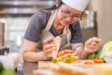Ahşap kesim tahtasının üzerinde salata, yumurta ve peynir katmanları olan taze bir sandviçi bir araya getiren şefin yakın plan fotoğrafı. Dilimlenmiş domatesler yakınlarda ve şef çizgili bir önlük giyiyor..