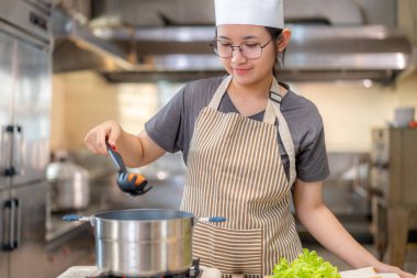 Çizgili önlüklü ve şapkalı genç bayan şef, ticari bir mutfakta kepçeyle tencereye yumurta ekliyor. Taze sebzeler ve ekmekle çevrili olarak, neşe ve profesyonellikle bir yemek hazırlıyor..