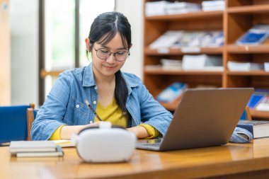 Kütüphanede dizüstü bilgisayarla çalışan odaklanmış genç bir kadın. Yanında bir VR kulaklığı geleneksel öğrenme ve eğitimde modern teknolojinin karışımını öneriyor..