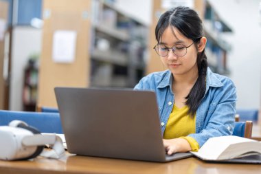 Kütüphanede dizüstü bilgisayarla çalışan odaklanmış genç bir kadın. Yanında bir VR kulaklığı geleneksel öğrenme ve eğitimde modern teknolojinin karışımını öneriyor..