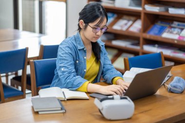 Kütüphanede dizüstü bilgisayarla çalışan odaklanmış genç bir kadın. Yanında bir VR kulaklığı geleneksel öğrenme ve eğitimde modern teknolojinin karışımını öneriyor..