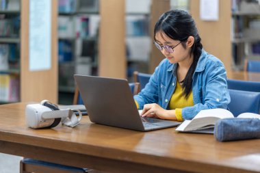 Kütüphanede dizüstü bilgisayarla çalışan odaklanmış genç bir kadın. Yanında bir VR kulaklığı geleneksel öğrenme ve eğitimde modern teknolojinin karışımını öneriyor..