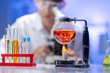 Laboratuvar tezgahındaki alevlerin üzerinde kırmızı bir sıvı ısıtılıyor. Arka planda, bulanık bir bilim adamı aktif bir araştırma ve geliştirme sahnesi yaratıyor..