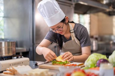 Profesyonel bir mutfakta, genç, mutlu bir Asyalı kadın şef, şef şapkası takıyor, titizlikle taze ve sağlıklı bir sandviç hazırlarken gülümsüyor. Yemek hazırlama, sağlıklı beslenme ya da mutfak yaratımı için ideal..