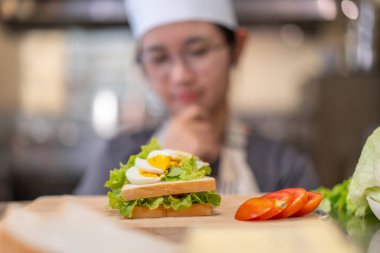 Üniformalı genç bir bayan şef hazırladığı sandviçi incelikle inceliyor, odaklanma, yaratıcılık ve mutfak ortamında aşçılık becerileri gösteriyor. Sandviçe odaklan..