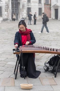 Venedik, İtalya - 22 Aralık 2022: Geleneksel Çin Guzheng enstrümanı çalan genç Asyalı kadın. Şehir meydanındaki sokak müzisyeni bir müzik aleti çalıyor. Dikey fotoğraf
