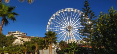Bulutları, yeşil palmiyeleri ve çam ağaçları olan mavi gökyüzünün arka planında büyük bir dönme dolap. Saranda, Arnavutluk.