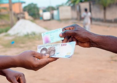 African hands exchanging Nigerian permanent voter's card with Naira notes, cash or money, depicting illegal activity of vote buying that often happens when it comes to elections in Nigeria