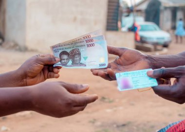African hands exchanging Nigerian permanent voter's card with Naira notes, cash or money, depicting illegal activity of vote buying that often happens when it comes to elections in Nigeria
