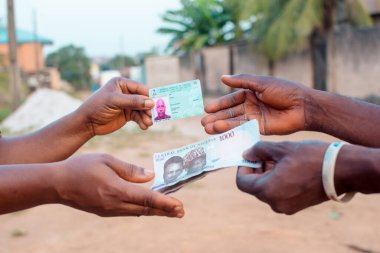 African hands exchanging Nigerian permanent voter's card with Naira notes, cash or money, depicting illegal activity of vote buying that often happens when it comes to elections in Nigeria