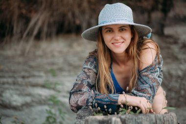 Uzun sarı saçlı, seksi mavi mayolu, küçük şapkalı ve mavi tişörtlü çekici ve genç bir kızın portresi doğada duruyor..