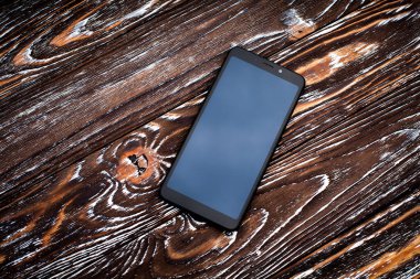 A black mobile phone with a dark display without an image lies on a dark wooden textured surface.