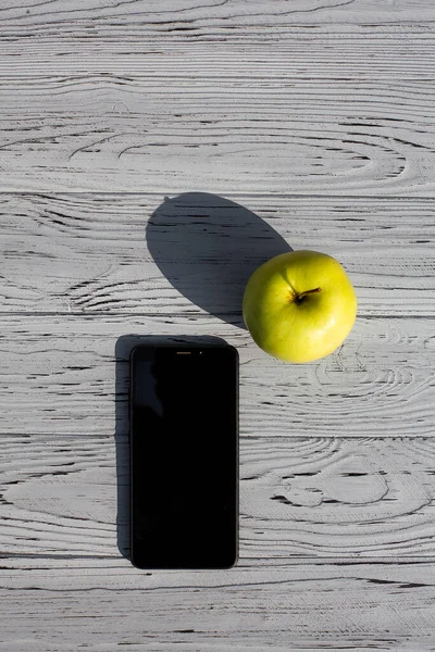 stock image A ripe, green apple lies near a mobile phone with a black display, on a light, textured, wooden table.