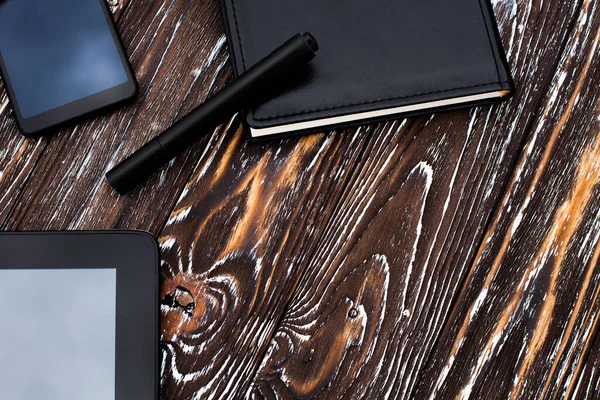 A black tablet, mobile phone and notebook with a leather cover and a pen lie on a dark wooden textured surface.