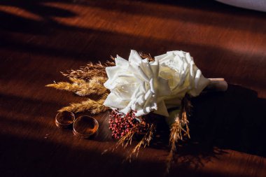 Golden wedding rings and the groom's boutonniere made of white roses and brown grass, lie on a wooden brown table.