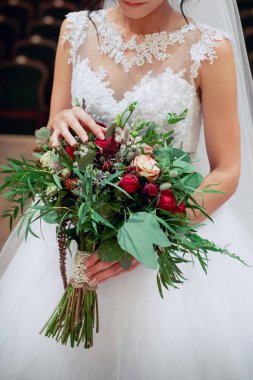 Beyaz bir gelinlik içinde genç bir gelin elinde yeşil yapraklı taze sarı ve kırmızı çiçeklerden bir buket tutuyor ve parmaklarıyla nazikçe dokunuyor.