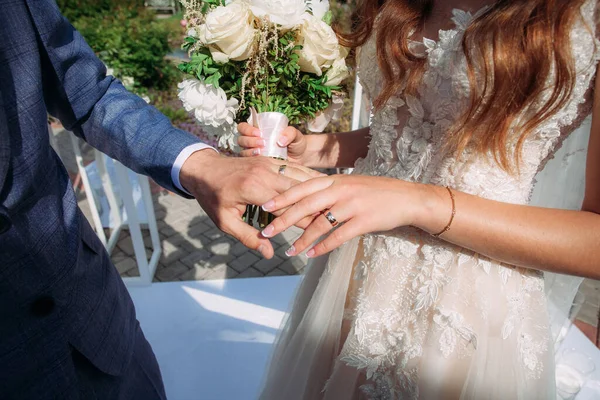 Gelin ve damat, parlak ve güneşli bir günde alyans takası seremonisinde birbirlerinin elini tutuyorlar. Gelinin elinde zarif bir buket beyaz ve sarı güller var..