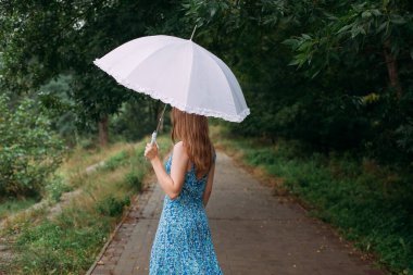 A slender young girl with blond long hair, in a blue summer dress, stands with a white umbrella in her hands, on a path in a forest park area in the rain.