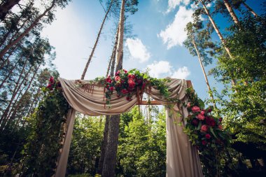 Ahşap bir üste, yeşil yapraklı ve bej perdeli taze kırmızı güllerin düğün töreni için şık bir düğün kemeri yaz boyunca ormanda durur..