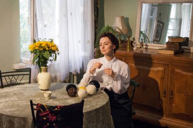 A stylish Ukrainian woman in vintage clothes sits at a table in a cozy apartment with retro furniture, holds knitting threads in her hands. There is a vase with sunflowers on the table.