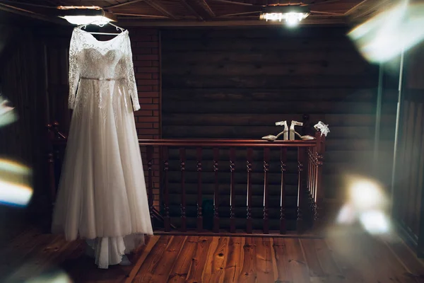 Art work. Wedding accessories, a white dress of the bride hangs on a chandelier, women's ivory-colored shoes, a white leg garter lie on a brown railing, in the interior of an apartment in a dark room.