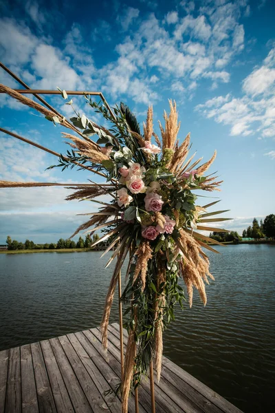 Art Deco wedding arch with pink roses and leaves on a wooden platform for the ceremony, against the background of the river, on a clear sunny day.