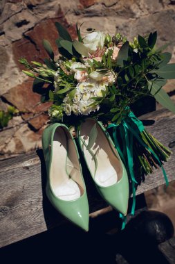 Beyaz ve turuncu çiçeklerin gelini için düğün buketi ve şık yeşil ayakkabılar güneşli bir günde sokakta bir bankta yatıyor..