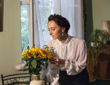 A stylish Ukrainian woman in vintage clothes, a black skirt and a white shirt, stands by the window in a cozy apartment with retro furniture. There is a vase with sunflowers on the table.