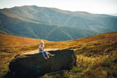 Mavi pantolonlu ve açık renk tişörtlü küçük bir kız sıcak bir yaz günü, dağlarda büyük bir taşın üzerinde oturur. Çimlerin, ağaçların ve bulutlu mavi gökyüzünün etrafında.