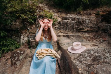 Mavi elbiseli genç bir kız güneşli bir günde dağlardaki kayalıklarda oturur ve yüzünü buruşturur. Yakınlarda pembe bir şapka ve bej bir çanta var..