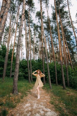 İnce, bej renkli bir elbise giyen genç bir kız açık güneşli bir günde ağaçların arkasında duruyor. Elinde hasır bir şapka tutuyor ve gülümsüyor..