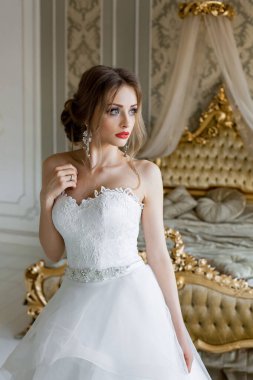 Şık makyajlı, beyaz bir gelinlik giymiş güzel bir gelin, düğün öncesi sabah şık bir iç mimariye sahip bir yatak odasında duruyor..