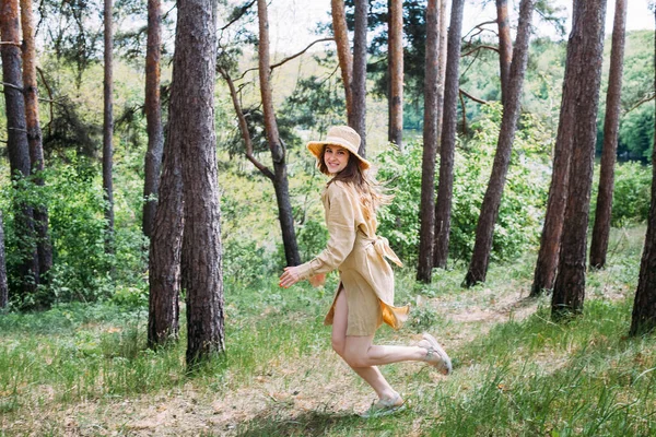 一个金发碧眼 头戴米黄色太阳衣 头戴一顶帽子的漂亮姑娘穿过森林 微笑着 图库图片