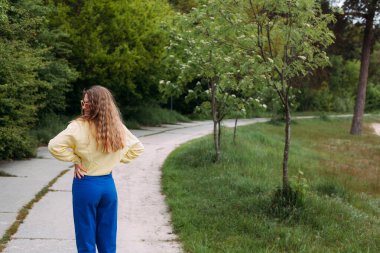 Sarı kıvırcık saçlı, mavi kot pantolonlu, sarı gömlekli, güneş gözlüklü, gülümseyen bir kız yaz mevsiminde resim parkında bir yolda duruyor..