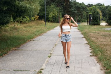 Uzun sarı saçlı, kısa seksi kot şortlu, beyaz bluzlu, güneş gözlüklü, parkta yürüyen ince bir kız..