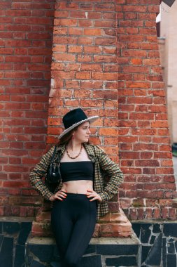A slender young girl with blond long hair, in black leggings, a tank top, a beige jacket and a hat, stands on the street near the wall of an old building.