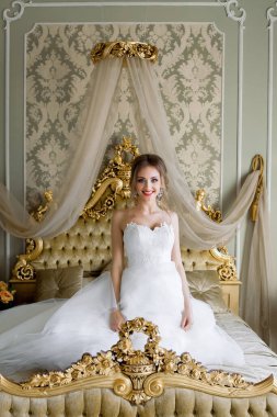 Beyaz gelinlikli güzel bir gelin, düğün öncesi sabah, şık bir iç dekorasyonu olan bir yatak odasında oturuyor..