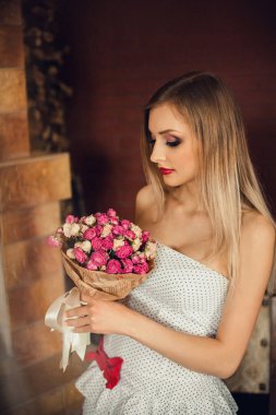 Parlak makyajlı, sarı saçlı, elbise giymiş muhteşem bir kız şömineli bir odada duruyor. Elinde güzel pembe ve beyaz güller tutuyordu..