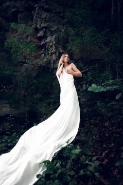 İnce yapılı, sarı saçlı güzel bir genç kız gün boyunca uzun beyaz bir kumaşa sarılı bir kayanın üzerinde duruyor..