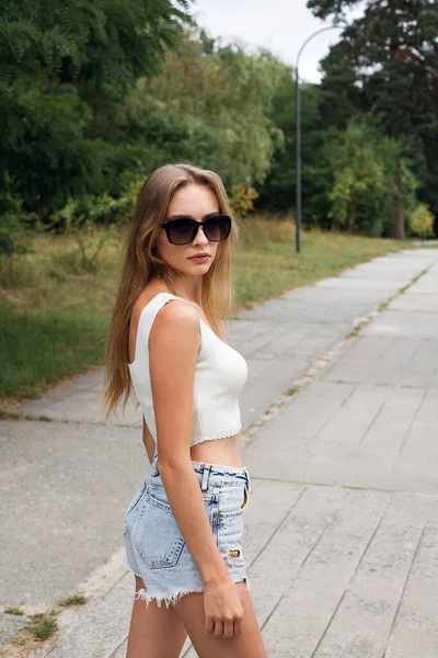 A young slender girl with long blond hair in short sexy denim shorts and a white top, in sunglasses, stands on a path in the park.