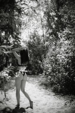 Art work. A black and white photograph of a girl in a black bathing suit stands alone in the forest, fixing her hair, on a sunny day.