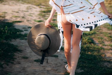 Güneşli bir yaz gününde elinde şapkayla tarlada yürüyen desenli tunik giymiş bir kız..