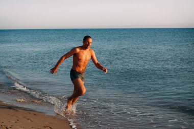 Mavi yüzme şortlu ince kaslı bir adam okyanusun kumlu kıyılarında koşuyor ve güneşli bir günde gülümsüyor..