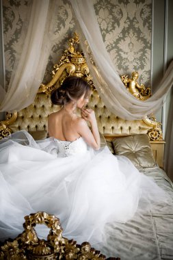 Şık makyajlı, beyaz bir gelinlik giymiş güzel bir gelin, düğün öncesi sabah şık bir iç dekorasyonla bir yatak odasında oturur..