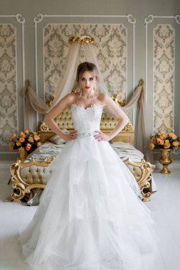 Şık makyajlı, beyaz bir gelinlik giymiş güzel bir gelin, düğün öncesi sabah şık bir iç mimariye sahip bir yatak odasında duruyor..