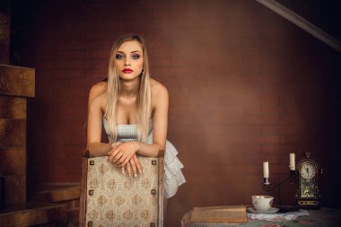 Parlak makyajlı, sarı saçlı, elbiseli, şömineli, masanın yanında duran muhteşem bir kız. Masanın üzerinde mum, saat, bardak ve kitap olan bir şamdan var..