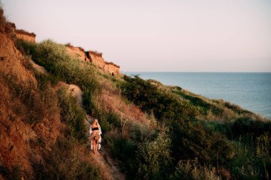 Siyah bikinili güzel bir kız ve elinde şapkası olan açık tenli bir pareo, açık güneşli bir günde, okyanus kenarındaki tepelerin arasında duruyor..